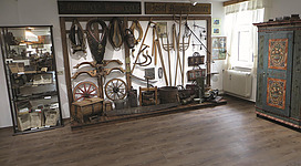 Ein Ausstellungsraum mit historischen Werkzeugen und landwirtschaftlichen Geräten. An der Wand sind verschiedene Objekte und eine verzierte Truhe zu sehen.