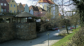 Seilergraben in Schärding: Eine Reihe historical Häuser mit traditioneller Architektur, eingebettet in eine grüne Umgebung mit einem Kirchturm im Hintergrund. Der Platz ist mit Pflastersteinen ausgelegt und durch einen Brunnen belebt. Autos sind auf einem kleinen Parkplatz abgestellt.