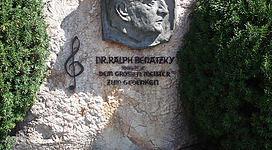 Ralph Benatzky Denkmal in St. Wolfgang im Salzkammergut: Das Bild zeigt ein Denkmal, bestehend aus einem Felsblock mit einem erhobenen Reliefprofil. Darunter befindet sich eine Inschrift, die eine prominente Persönlichkeit zu würdigen scheint. Das Denkmal ist von üppiger Vegetation umgeben, was eine angenehme und ruhige Umgebung schafft.