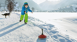 Ein Junge spielt im Schnee und rollt eine rote Kugel. Im Hintergrund sind Berge und ein Hund zu sehen.