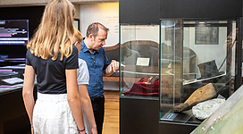 Riesenmuseum in Lengau: In dem Bild sind zwei Personen zu sehen, die sich gemeinsam eine Ausstellung in einem Museum ansehen. Sie stehen vor einer Vitrine, in der verschiedene alte Gegenstände und Artefakte ausgestellt sind. Der Mann betrachtet die Exponate mit Interesse, während die Frau neben ihm steht und den Ausstellungsraum erkundet.