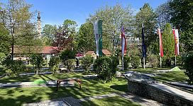 ASKÖ Minigolfplatz in Braunau am Inn: Ein Park mit gepflegten Grünanlagen, Bäumen und Büschen, in dem sich Denkmäler und Fahnen in der Sonne abheben. Es gibt einen Brunnen und Bänke für Besucher. Die Umgebung wirkt friedlich und einladend.