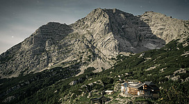 Eine einsame Berghütte liegt in einer grünen Landschaft unterhalb einer beeindruckenden Bergkette. Der Himmel ist klar und die Umgebung ist geprägt von Felsen und Wäldern.