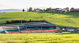 Union Tennisclub Pfaffing-Vöcklamarkt in Vöcklamarkt: Das Bild zeigt eine Sportanlage mit einem Tennisplatz inmitten einer grünen Landschaft. In der Ferne sind Gebäude zu sehen, die eine ländliche Umgebung andeuten.