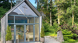Ein modernes, kubisches Gebäude mit einem Kreuz auf dem Dach steht im Wald. Der Garten ist mit grünen Pflanzen und Steinen gestaltet.