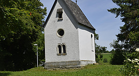 Eine kleine weiße Kapelle steht inmitten von grünen Wiesen und Bäumen. Der Himmel ist klar und die Umgebung wirkt ruhig und friedlich.