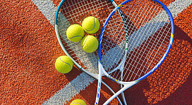Zwei Tennisschläger liegen auf einem Tennisplatz zusammen mit mehreren Tennisbällen. Der Untergrund ist orangefarben und in gutem Zustand.