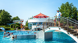 Freibad Leonding in Leonding: Eine große Freiluftanlage mit einem großen Pool, an dessen Rand mehrere Personen planschen und sich erfrischen. Das Gelände ist umgeben von grünen Bäumen und einer roten überdachten Struktur, vermutlich eine Art Pavillon.