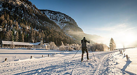 Ein Skifahrer gleitet durch eine verschneite Winterlandschaft. Im Hintergrund sind Berge und Bäume zu sehen, während die Sonne aufgeht.