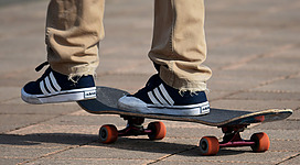 Skaterplatz in Schörfling am Attersee: Das Bild zeigt die Füße einer Person, die auf einem Skateboard steht. Das Skateboard ist schwarz mit weißen Streifen, und die Person trägt blaue Schuhe und hellbraune Hosen.