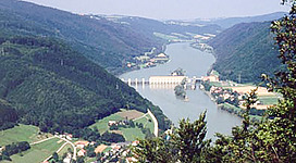 Europaschutzgebiet Oberes Donau- und Aschachtal in Aschach an der Donau: Eine malerische Landschaft mit einem großen Stausee oder Fluss, umgeben von bewaldeten Bergen. In der Mitte des Bildes befindet sich ein imposantes Gebäude, vermutlich ein Wasserkraftwerk oder eine Talsperre. Kleine Ortschaften sind auf den umliegenden Anhöhen zu erkennen. Die Szene vermittelt einen friedlichen und ruhigen Eindruck.