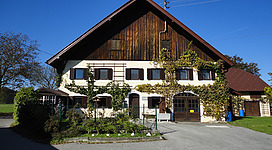Moorschmiede Gerlham in Seewalchen am Attersee: Das Bild zeigt ein traditionelles, hölzernes Gebäude mit weißen Fassadenteilen und einem steilen Dach. Es ist von Grünflächen und einzelnen Bäumen umgeben und wirkt gemütlich und einladend.