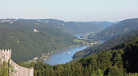 Donaublick Flenkental in St. Aegidi: Eine malerische Landschaft mit einem See, umgeben von bewaldeten Bergen und Hügeln. Kleine Häuser und Gebäude sind im Tal verteilt. Eine Straße windet sich durch die Landschaft und ein Boot ist auf dem See zu sehen.