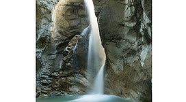 Burggrabenklamm in Steinbach am Attersee: Das Bild zeigt einen malerischen Wasserfall, der aus einer felsigen Schlucht herunterstürzt. Das türkisfarbene Wasser sammelt sich in einem ruhigen Becken am Fuße des Wasserfalls, umgeben von felsigen Wänden und Vegetation.
