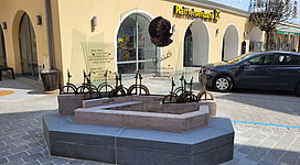 Rudi Nierlich Denkmal in St. Wolfgang im Salzkammergut: Das Bild zeigt ein Gebäude mit einem markanten Eingang und einem Brunnen davor. Der Brunnen hat mehrere aufragende Metallstangen, die an eine Stahlskulptur erinnern. Außerdem ist ein Auto auf der Straße vor dem Gebäude zu sehen.