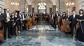 Ein großes Orchester steht in eleganter Kleidung auf einer Bühne. Die Musiker halten verschiedene Instrumente wie Geigen und Cellos.