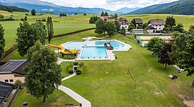 Ein Schwimmbad mit Rutschen und mehreren Pools in einer grünen Landschaft. Im Hintergrund sind Berge und einige Häuser sichtbar.