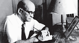 Leo Perutz Denkmal in St. Wolfgang im Salzkammergut: Das Bild zeigt einen älteren Herrn in Anzug und Krawatte, der konzentriert vor einem Schreibtisch sitzt und in Papiere vertieft ist. Er hat ein ernsthaftes Gesichtsausdruck und trägt eine Brille.