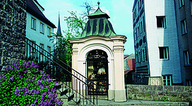 Nepomukkapelle in Braunau am Inn: Ein schönes historisches Gebäude mit einer reich verzierten Toreinfahrt, umgeben von blühenden Pflanzen. Der Turm des Gebäudes ragt in den blauen Himmel. Das Ensemble wirkt sehr malerisch und einladend.