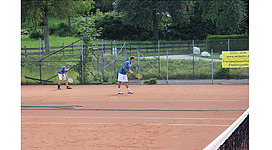 Tennisclub Schörfling in Schörfling am Attersee: Das Bild zeigt zwei Männer, die auf einem Tennisplatz spielen. Der Platz ist mit einer Rasenfläche und Umrandungen versehen, umgeben von dichter, grüner Vegetation. Es sieht nach einem Erholungsort oder einer Sportanlage aus.