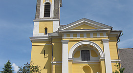 Mühlviertler Dom in Niederkappel: Eine große, gelblich gestrichene Kirche mit einem hohen Glockenturm und einer Uhr an der Fassade. Vor dem Haupteingang der Kirche führen Stufen hinauf und es gibt einen kleinen Garten mit Bäumen. Insgesamt zeigt das Bild ein traditionelles, historisches Kirchengebäude in einer ländlichen Umgebung.