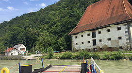 Getreidespeicher in Kirchberg ob der Donau: Eine malerische Landschaft mit einem alten, traditionell aussehenden Gebäude, das am Ufer eines Flusses steht. Vor dem Gebäude befinden sich einige Boote und ein hölzerner Steg. Die Umgebung ist von üppigem Grün, Bäumen und Bergen geprägt, die eine idyllische und friedliche Atmosphäre vermitteln.