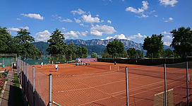 Tennisclub Nußdorf in Nußdorf am Attersee: Das Bild zeigt einen Tennisplatz mit roter Aschebahn, umgeben von Bäumen, Sträuchern und Bergen im Hintergrund. Mehrere Personen sind beim Tennisspielen zu sehen.