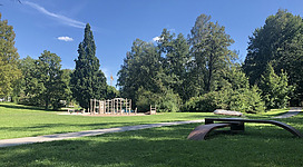 Stadtpark Ried im Innkreis in Ried im Innkreis: Eine weitläufige, grüne Parklandschaft mit hohen Bäumen und Wiesen. Im Vordergrund befinden sich einige Bänke und ein Spielplatz mit Klettergerüsten. Der Himmel ist strahlend blau mit vereinzelten Wolken. Eine friedliche und einladende Umgebung.