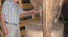 Steublmühle Gramastetten in Gramastetten: Ein älterer Mann, der in einem alten Schuppen steht und auf einen großen, runden Gegenstand an der Wand deutet. Der Schuppen ist aus Holz und enthält viele verschiedene Holzmaterialien und Werkzeuge. Der Mann trägt eine Kappe und karierte Kleidung.