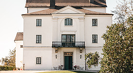 Schloss Wolfsegg in Wolfsegg am Hausruck: Ein historisches, prächtiges Herrenhaus mit traditioneller Architektur und einem ansprechenden Garten. Das Gebäude weist einen imposanten Haupteingang, weißgetünchte Fassaden und ein markantes Dach auf. Davor erstreckt sich eine gepflegte Rasenfläche, die von Büschen und Bäumen umgeben ist.