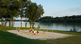 Beach-Volleyballplatz Au/Donau in Naarn im Machlande: An idyllische Szene mit Blick auf einen ruhigen See, umrahmt von üppiger, grüner Vegetation. Auf einem Sandstrand befindet sich ein Volleyballnetz, an dem mehrere Menschen spielen. Ein friedlicher Ort, der zu sportlichen Aktivitäten und Erholung einlädt.