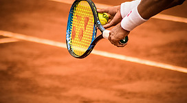 Ein Tennisschläger hält einen Tennisball. Der Hintergrund zeigt einen roten Sandplatz.