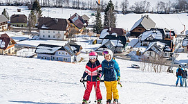 Zwei Kinder in Skibekleidung stehen auf einem schneebedeckten Hang. Im Hintergrund sind eine Ski-Region und kleine Häuser zu sehen.