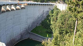 Ein großer Damm umgeben von Bäumen und Bergen. Darunter befindet sich ein grünes Wasserbecken.