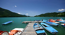Ein malerischer Steg am Wasser mit mehreren bunten Booten. Im Hintergrund sind sanfte Hügel und ein klarer blauer Himmel zu sehen.