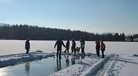 Eine Gruppe von Menschen steht auf einem gefrorenen See, umgeben von Schnee. Der Himmel ist klar und sonnig.