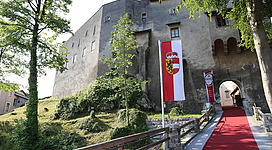 Eine alte Burg mit einer roten Teppichauffahrt und einer Flagge. Bäume umgeben das Gebäude und der Himmel ist klar.