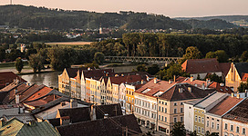 Walcher- und Färbergraben in Braunau am Inn: Eine malerische Altstadt mit farbenfrohen, mehrstöckigen Gebäuden, die sich an einem Fluss erstrecken. In der Ferne ragen Berge auf, die eine idyllische Kulisse bilden. Die Häuser sind größtenteils mit roten Ziegeldächern bedeckt, was dem Stadtbild einen traditionellen Charakter verleiht. Dazwischen erheben sich einige Kirchtürme, die das Panorama akzentuieren.