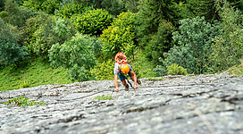 Eine Person klettert an einer steilen Felswand. Im Hintergrund sind grüne Bäume und eine üppige Landschaft zu sehen.