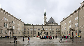 Domplatz (Cathedral Square) in the historic centre of the city of Salzburg, as seen from the cathedral. The square is surrounded by a part of Salzburg Residence. In the center: Wallistrakt which houses lecture halls of the University of Salzburg. In the background: Franciscan church.