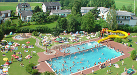 Erlebnisfreibad Lembach in Lembach im Mühlkreis: Ein kommerzielles Freizeitzentrum mit einem großen Außenpool, von dem aus der Blick auf die umgebende Landschaft mit Häusern und Wald fällt. Viele Menschen nutzen den Pool und die umliegenden Liegewiesen für Erholung und Entspannung.