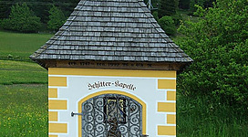 Eine kleine Kapelle mit einem spitzen Dach steht in einer grünen Umgebung. Vor der Kapelle befindet sich ein dekoratives Gitterfenster mit Blumen.