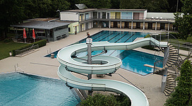 Freizeitpark Vöcklabruck in Vöcklabruck: Ein Resort-Komplex mit mehreren Gebäuden und Schwimmbecken. Die Architektur ist modern, mit bunten Fassaden und geschwungenen Linien. Die Anlage ist von üppigen Grünanlagen umgeben, darunter hohe Bäume. Eine Rutsche führt vom höheren in ein niedrigeres Becken. Insgesamt vermittelt das Bild einen einladenden und erholsamen Eindruck.