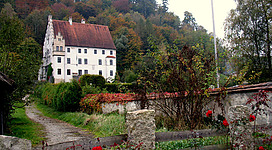Schloss Wanghausen in Hochburg-Ach: Ein altes, mehrstöckiges Gebäude mit einem roten Ziegeldach, umgeben von herbstlich gefärbten Bäumen und einer Steinmauer. Im Vordergrund befinden sich einige rote Blumen auf dem Rasen. Die Umgebung wirkt idyllisch und friedlich.