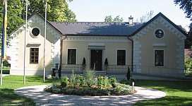Heimatmuseum Schwanenstadt in Schwanenstadt: Ein zweistöckiges Gebäude mit klassischer Architektur, umgeben von Bäumen und einem gepflegten Garten. Die Fassade ist hell gestrichen und weist verschiedene Fensterformen auf, darunter große Rundbogenfenster. Das Dach ist mit dunklen Ziegeln gedeckt. Ein gepflasterter Weg führt zum Eingang des Gebäudes.