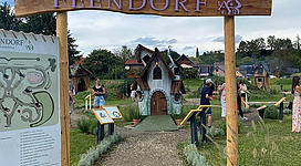 Ein schöner Eingang zu Feendorf mit einem Holzschild. Im Hintergrund sind kleine, charmante Häuschen und Grünflächen zu sehen.