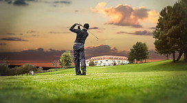 Ein Golfer schlägt einen Ball auf einem gepflegten Golfplatz bei Sonnenuntergang. Im Hintergrund sind Bäume und ein Gebäude sichtbar.