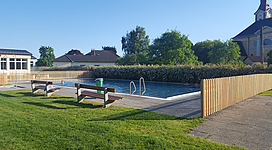 Freibad Niederkappel in Niederkappel: Eine idyllische Umgebung mit einem schmalen Schwimmbecken, umgeben von einem Rasen und umstanden von Gebäuden mit traditioneller Architektur. Die Spiegelung der umgebenden Vegetation und Strukturen im Wasser verleiht der Szene einen friedlichen und harmonischen Eindruck.