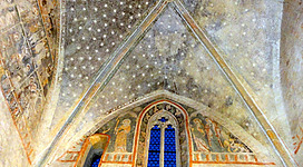 Frauenturm mit Johanniterkapelle in Enns: Eine beeindruckende gewölbte Decke mit einem Muster aus Sternen überzieht den Raum. Bunte, detailliert gestaltete Wandgemälde säumen die Wände, einschließlich einer markanten, blau gefärbten Fensterfront.