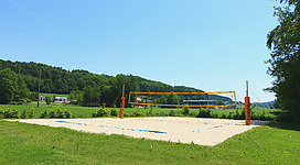 Beach-Volleyballplatz in Maria Schmolln: Eine grüne Wiese mit einem Volleyballplatz, umgeben von Bäumen und einer bergigen Landschaft. Im Vordergrund befindet sich ein Volleyballfeld mit Netz und Begrenzungslinien.